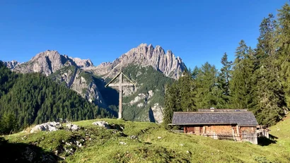 Holzkreuz und Berghütte vor schroffen Bergen und blauem Himmel