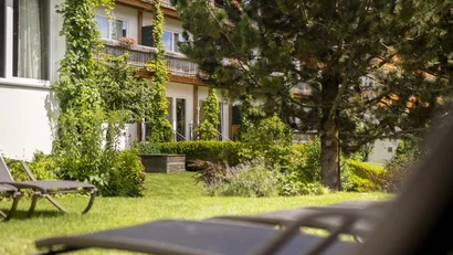 Sonniger Garten mit grünen Pflanzen und Liegestühlen vor einem Wohngebäude