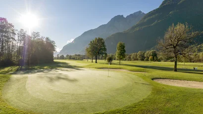 Golfplatz mit Bäumen und Bergen im Hintergrund bei sonnigem Wetter