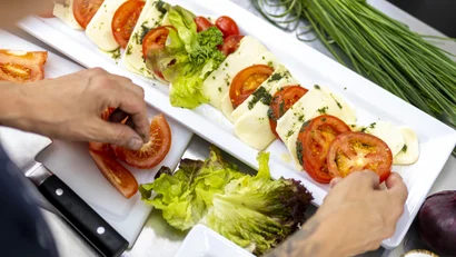 Person richtet Tomatenscheiben und Käse mit Kräutern auf weißem Teller an